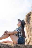 belle femme en chemise à carreaux et chapeau de cowboy reposant sur une botte de foin photo