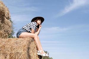 belle femme en chemise à carreaux et chapeau de cowboy reposant sur une botte de foin sur fond de ciel photo