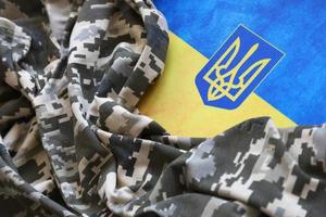 sumy, ukraine - 20 mars 2022 drapeau ukrainien et armoiries avec tissu à texture de camouflage pixélisé. tissu avec motif camouflage en formes de pixels gris, marron et vert avec le signe du trident ukrainien photo