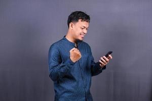 heureux jeune homme en chemise bleue posant isolé sur fond gris portrait en studio. concept de mode de vie des gens. maquette de l'espace de copie. tenir le téléphone portable, faire le geste du gagnant photo