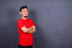 portrait d'un homme asiatique séduisant en t-shirt rouge debout avec les bras croisés isolé sur fond gris photo