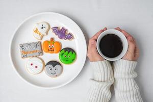 main de femme tenant une tasse de café en mangeant des biscuits d'halloween drôles. bonne fête d'halloween, astuce ou menace, bonjour octobre, automne automne, concept traditionnel, fête et vacances photo