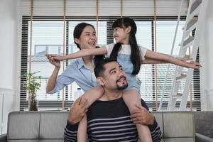 famille thaïlandaise asiatique ensemble, papa joue et taquine avec sa fille et sa maman en portant et en tenant une fille sur les épaules dans le salon de la maison, des moments de loisirs heureux, un week-end agréable, un style de vie domestique bien-être. photo