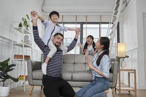 famille thaïlandaise asiatique ensemble, papa joue et taquine avec les enfants et maman en portant et en tenant son fils sur les épaules dans le salon de la maison, des loisirs heureux, un week-end agréable, un style de vie domestique bien-être. photo