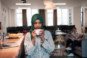 femme d'affaires musulmane africaine portant un hijab vert buvant du thé tout en travaillant sur un ordinateur portable dans un espace de détente au bureau de démarrage moderne à aire ouverte. photo
