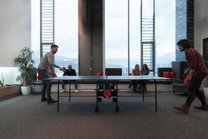 deux jeunes hommes d'affaires en démarrage jouant au tennis de table dans un espace de bureau créatif moderne, un groupe de personnes se réunit et réfléchit en arrière-plan photo