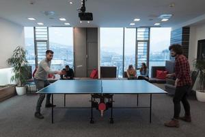 deux jeunes hommes d'affaires en démarrage jouant au tennis de ping-pong dans un espace de bureau créatif moderne, un groupe de personnes se réunit et réfléchit en arrière-plan photo