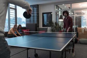 deux jeunes hommes d'affaires en démarrage jouant au tennis de ping-pong dans un espace de bureau créatif moderne, un groupe de personnes se réunit et réfléchit en arrière-plan photo