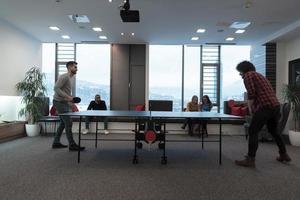 deux jeunes hommes d'affaires en démarrage jouant au tennis de table dans un espace de bureau créatif moderne, un groupe de personnes se réunit et réfléchit en arrière-plan photo