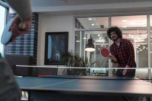 deux jeunes hommes d'affaires en démarrage jouant au tennis de ping-pong dans un espace de bureau créatif moderne, un groupe de personnes se réunit et réfléchit en arrière-plan photo