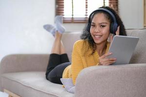 jeune femme africaine se détendant à la maison allongée sur un canapé et écoutant de la musique sur une tablette portant des écouteurs. fille relaxante sur le canapé d'une maison confortable photo