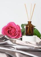 Diffuseur d'arômes pour la maison avec des bâtons de roseau, parfum de fleur de rose. maison cosy, ambiance romantique, draps en satin de soie. fleur rose rose à côté d'une bouteille en verre avec de l'huile parfumée. photo