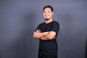 souriant bel homme asiatique en t-shirt noir debout avec les bras croisés isolé sur fond gris photo