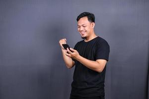 heureux homme asiatique tenant un smartphone et remportant le prix. isolé sur fond gris photo