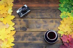 une tasse de thé et un vieil appareil photo parmi un ensemble de feuilles d'automne tombées jaunissantes sur une surface de fond de planches en bois naturel de couleur marron foncé