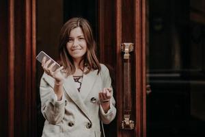 élégante femme magnifique en imperméable, tient un téléphone portable, attend un appel, pose à l'extérieur près des portes, va au travail, se sent heureuse, met à jour les informations sur le site Web, connectée à Internet haut débit photo