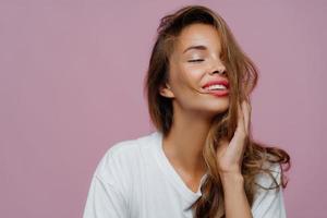 belle femme détendue aux longs cheveux blonds, garde les yeux fermés du plaisir, sourit positivement, aime être photographiée, pose devant la caméra, montre que sa beauté a du maquillage isolé sur le mur violet du studio photo