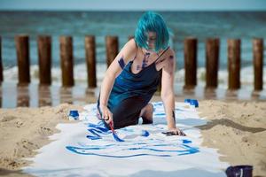 Artiste de performance artistique femme aux cheveux bleus enduite de peintures à la gouache sur une grande toile sur la plage photo