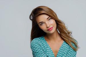 photo d'une jolie jeune femme incline la tête, a une expression ravie, porte du maquillage, a de longs cheveux ondulés, vêtue d'une chemise à pois, regarde agréablement la caméra, isolée sur un mur blanc. portrait beauté