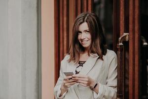 brune gaie charmante femme envoie une lettre par e-mail, utilise un téléphone portable et une connexion Internet gratuite, vêtue d'une veste blanche, se tient près des portes du bâtiment. personnes, mode de vie, concept technologique photo