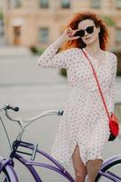 une femme aux cheveux rouges réfléchie concentrée sur la distance, garde la main sur le cadre des lunettes de soleil, pose près du vélo prend une pause après avoir parcouru une longue distance porte une robe d'été pose en plein air sur fond flou photo