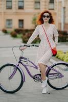 photo verticale d'un beau modèle féminin aux cheveux roux se repose après avoir parcouru une longue distance à vélo, porte des lunettes de soleil, une robe d'été et des baskets blanches, aime le sport et explore les rues de la ville