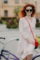 photo d'une femme heureuse aux cheveux rouges avec un doux sourire, passe son temps libre à faire du vélo dans les rues de la ville pendant une journée ensoleillée et venteuse, porte une robe d'été élégante pose sur un arrière-plan flou de la ville. tir en plein air