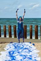 femme heureuse artiste de performance en robe bleue enduite de peintures à la gouache bleue dansant sur la plage de sable photo