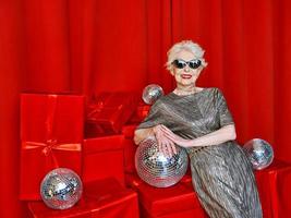 femme élégante et élégante senior avec vin de boule disco sur fond rouge. fête, mode, célébration, concept anti-âge photo