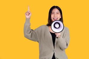 heureuse belle femme asiatique isolée sur fond jaune. elle crie à travers un mégaphone et pointe le côté photo