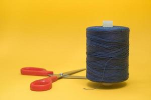 bobine de fil avec des ciseaux. une grande bobine de fil bleu se dresse sur un fond jaune. des ciseaux rouges se trouvent à proximité.