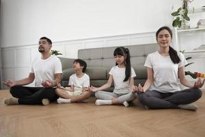 une famille thaïlandaise asiatique heureuse, des parents et des enfants s'assoient sur le sol du salon, ferment les yeux ensemble, méditent tranquillement en position du lotus et pratiquent un yoga sain, un week-end de style de vie à la maison. photo