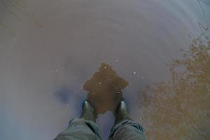 jambes dans des bottes en caoutchouc vert et un pantalon vert debout dans une flaque d'eau brune sale - vue à la première personne photo