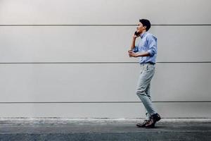 heureux jeune homme d'affaires en tenue décontractée utilisant un téléphone portable tout en marchant près du mur du bâtiment urbain. mode de vie des gens modernes. vue de côté. toute la longueur photo