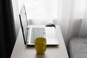 concept de bureau à domicile. travail à domicile dans le salon. ordinateur portable et tasse de boisson chaude sur une table en bois près du canapé et de la fenêtre. photo