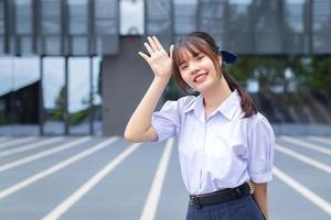 jolie lycéenne asiatique en uniforme scolaire se dresse et sourit joyeusement tout en toute confiance avec le bâtiment gris en arrière-plan. photo