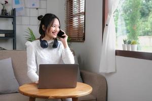 charmante jeune femme asiatique se détend dans le salon en appréciant de parler avec son collègue sur son téléphone tout en travaillant à domicile. photo