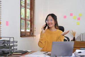 une femme d'affaires asiatique attrayante et souriante parle sur un téléphone portable avec un client avec un ordinateur portable au bureau. photo