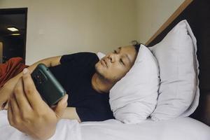 jeune homme fatigué vérifiant son téléphone en position couchée dans son lit photo
