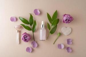 un sérum pour le visage ou une huile essentielle dans un flacon compte-gouttes rose posé sur un fond beige avec des pétales de rose et des feuilles vertes autour photo