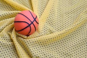 petit basket-ball en caoutchouc orange se trouve sur une texture et un fond de tissu de vêtements de maillot de sport jaune avec de nombreux plis photo