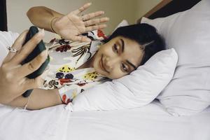 excitée joyeuse jeune femme asiatique faisant un appel vidéo ou un selfie tourné sur un téléphone portable en agitant la salutation en position couchée sur le lit photo