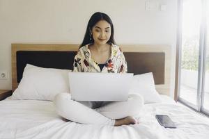 heureuse jeune femme travaillant depuis le lit. tenez l'ordinateur portable sur les genoux et tapez sur le clavier. photo