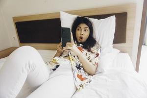 jeune femme choquée vérifiant son téléphone en position couchée dans son lit photo