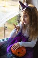 une fille en costume de sorcière sur le rebord de la fenêtre près de la fenêtre joue avec une toile d'araignée et une araignée, décorant la maison pour les vacances d'halloween. paysages terribles, peur et horreur, citrouilles photo
