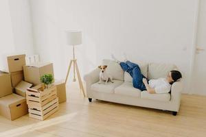 la locataire est allongée sur un canapé blanc confortable, lève les jambes croisées, pose avec son chien préféré, a un nouveau logement dans le salon, profite de la journée de réinstallation, se sent détendue. nouveau concept de maison et de déménagement photo