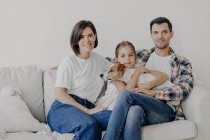 famille amicale pose ensemble au canapé, aime l'atmosphère domestique. le père, la mère, leur petite fille et leur chien de race passent le week-end à la maison, posent dans le salon, ont des expressions de visage heureux photo