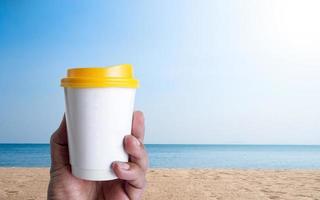 main d'homme tenant une tasse de café au poivre blanc couvercle jaune à emporter, rempli de café noir chaud sans lait de sucre, boisson prête, rafraîchissante. arôme éveillé frais au travail placé profiter de la mer plage ciel bleu soleil. photo