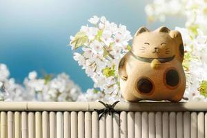 japon maneki neko ou chat faisant signe debout sur une clôture en bambou au-dessus de la fleur de sakura en fleurs japonaises. mascotte de chance, de fortune et d'argent photo