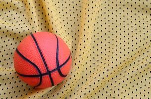 petit basket-ball en caoutchouc orange se trouve sur une texture et un fond de tissu de vêtements de maillot de sport jaune avec de nombreux plis photo